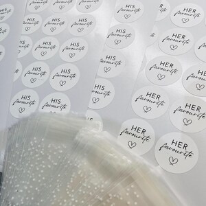 May include: A sheet of white stickers with black text that reads "His favourite" and "Her favourite" with a small heart symbol. The stickers are arranged in rows and columns.