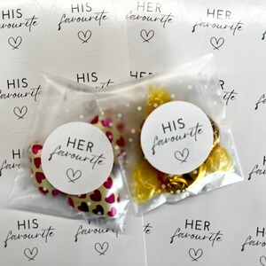 May include: Two clear plastic bags filled with small wrapped sweets. One bag has a white circle sticker that says "HIS favourite" with a heart. The other bag has a white circle sticker that says "HER favourite" with a heart. There are several white circle stickers with the same text on a white background.
