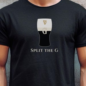 Pode incluir: T-shirt preto com um gráfico de um copo de stout, uma harpa e a letra "G". O texto "SPLIT THE G" está abaixo do gráfico em uma fonte serifada. A camisa é feita de um material macio.