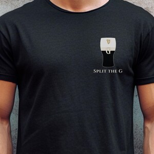 以下が含まれることがあります： 黒いTシャツ。スタウトのパイントグラスのグラフィックと「Split the G」の文字がプリントされています。パイントグラスのデザインは、黒と白で、金色のハープのシンボルが付いています。Tシャツはクルーネックです。