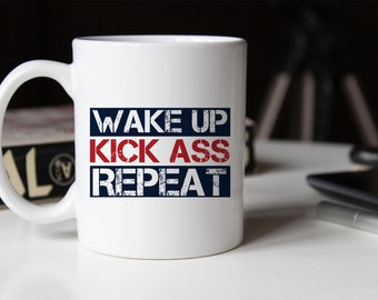 Könnte beinhalten: Ein weißer Keramikbecher mit dem Text WAKE UP in Dunkelblau, KICK ASS in Rot und REPEAT in Dunkelblau. Die Schrift hat eine abgenutzte, verwitterte Textur. Der Becher hat einen Standardhenkel auf der linken Seite und einen glatten, zylindrischen Körper.