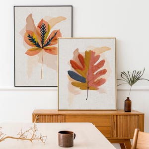 Op de afbeelding: Twee ingelijste kunstprints met bladeren. Eén toont een groot oranje en geel blad met donkerblauwe accenten, de andere een vergelijkbaar ontwerp met rode, gele en blauwe bladeren. De kunst wordt op een witte achtergrond weergegeven.