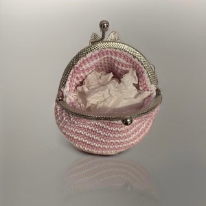 Può includere: Un piccolo portamonete all'uncinetto aperto, a righe rosa e bianche. La borsa ha una chiusura in metallo color argento ed è riempita di carta velina bianca. La borsa è su una superficie riflettente.