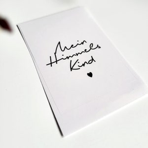 May include: A white card with the text "Mein Himmels Kind" in black script, accompanied by a small black heart. The card is rectangular and appears to be made of paper. The background is white.