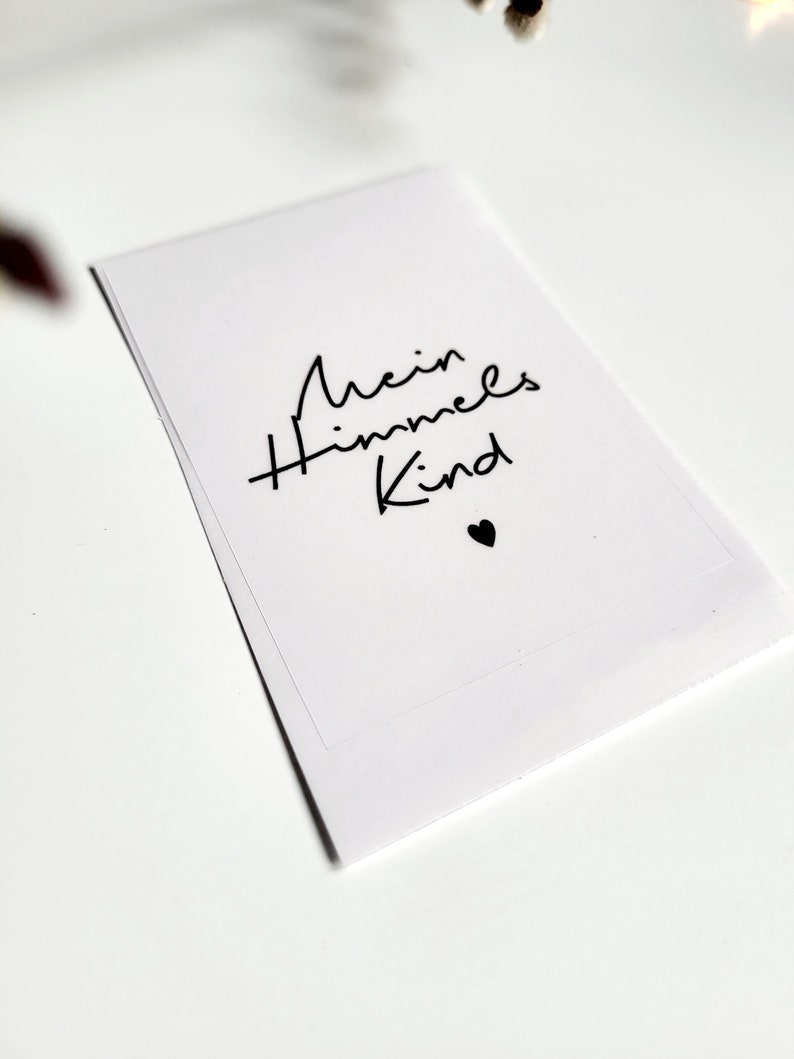 May include: A white card with the text "Mein Himmels Kind" in black script, accompanied by a small black heart. The card is rectangular and appears to be made of paper. The background is white.