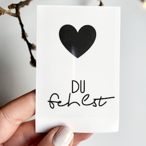 Könnte beinhalten: Eine weiße Karte mit einem schwarzen Herzen und dem Text "DU fehlest" in schwarzer Kursivschrift.