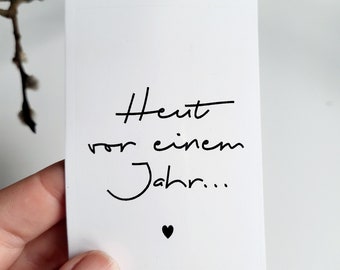Può includere: Un biglietto bianco con testo nero scritto a mano che recita "Heut vor einem Jahr..." e un piccolo cuore nero. Il biglietto è tenuto in mano, con un ramo sfocato sullo sfondo.