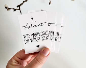Può includere: Una mano tiene in mano una pila di carte bianche con testo scritto a mano in nero. La carta in alto recita "1. Advent t-" e "WIR WÜNSCHTEN TEN EN DU WÄRST HIER! R! R!" è visibile. Un piccolo cuore nero è stampato sulla carta. Un ramo con gemme è sullo sfondo.