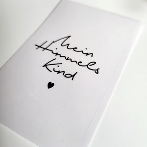 May include: A white card with the words "Mein Himmels Kind" in black script, accompanied by a small black heart. The card is rectangular and appears to be made of paper or cardstock.