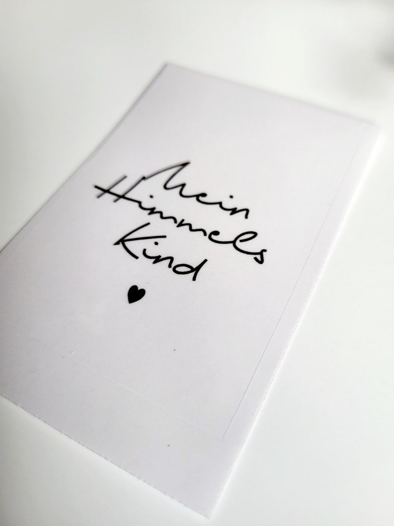 May include: A white card with the words "Mein Himmels Kind" in black script, accompanied by a small black heart. The card is rectangular and appears to be made of paper or cardstock.