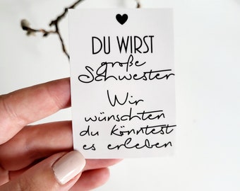 Può includere: Un biglietto bianco con testo nero che recita "Du wirst große Schwester. Wir wünschen du könntest es erleben." Il biglietto ha un piccolo cuore nero nell'angolo in alto a sinistra.