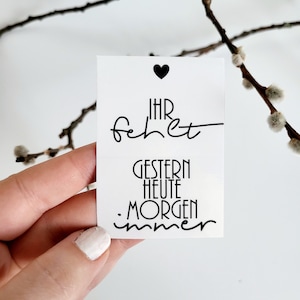 Könnte beinhalten: Eine weiße Karte mit schwarzem Text und einem schwarzen Herzsymbol. Die Karte zeigt die Worte "IHR fehet" über "GESTERN HEUTE MORGEN immer". Die Karte wird in einer Hand gehalten, mit einem verschwommenen Hintergrund aus Ästen.