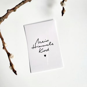 May include: A white card with the text "Mein Himmels Kind" in black script, with a small black heart below the text. A brown branch with small, fluffy buds is in the upper left corner. The background is white.