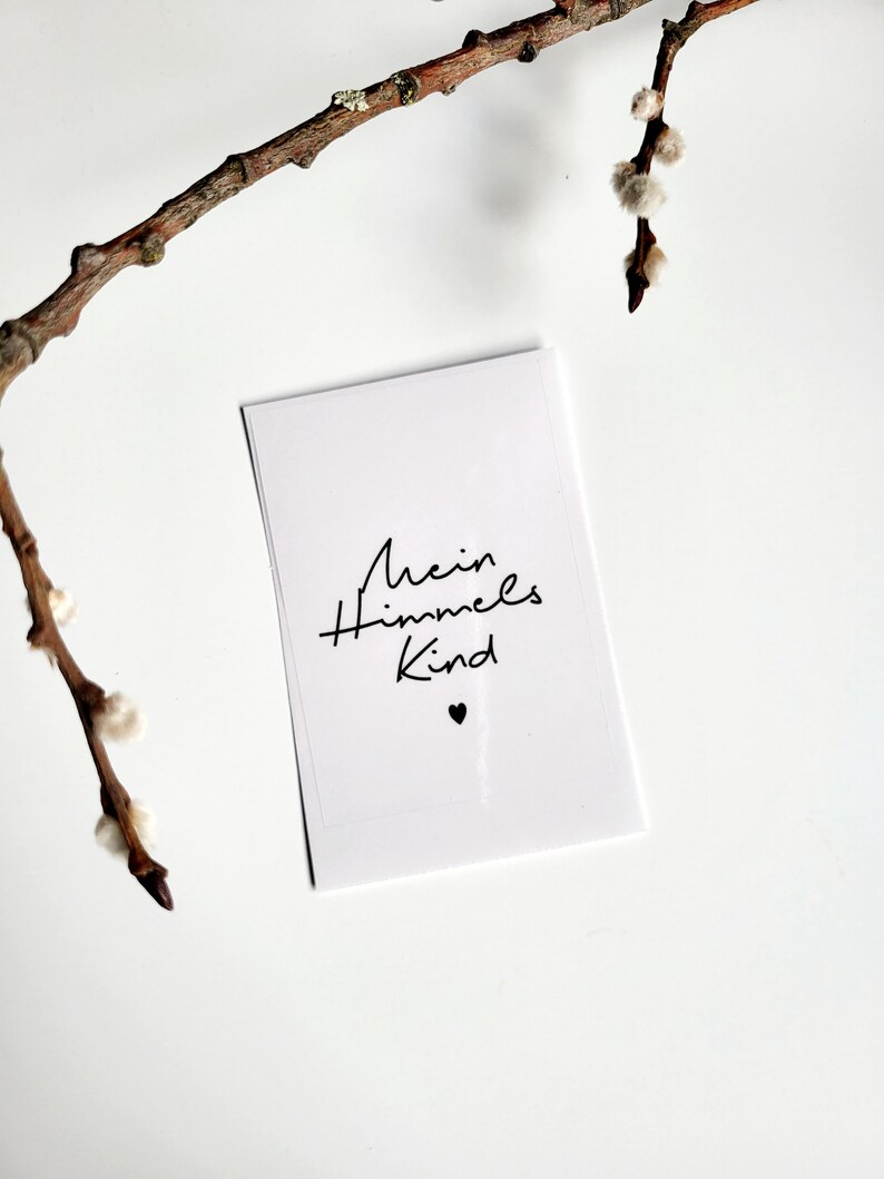May include: A white card with the text "Mein Himmels Kind" in black script, with a small black heart below the text. A brown branch with small, fluffy buds is in the upper left corner. The background is white.