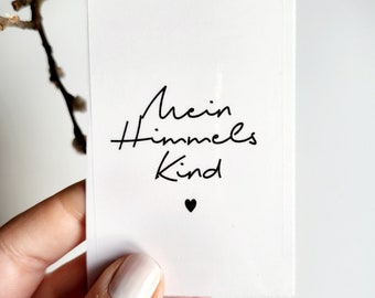 Può includere: Una carta bianca con la scritta tedesca "Mein Himmels Kind" in corsivo nero, con un piccolo cuore nero sotto. La carta è tenuta da una persona con lo smalto chiaro, con un ramo sfocato sullo sfondo.