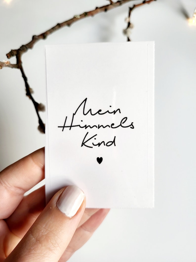 May include: A white card with the German text "Mein Himmels Kind" in black script, with a small black heart below the text. The card is held by a person with light-colored nail polish, with a blurred branch in the background.