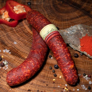 Könnte beinhalten: Zwei Würste von rötlich-brauner Farbe mit einem Etikett, zusammen mit roten Paprikascheiben, Pfefferkörnern und rotem Paprikapulver auf einer Holzoberfläche. Die Wurst hat eine strukturierte Oberfläche.