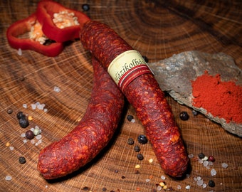 May include: Two links of reddish-brown sausage with a label, alongside red bell pepper slices, peppercorns, and red paprika powder on a wooden surface. The sausage has a textured surface.