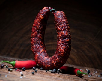 May include: A ring of smoked sausage with a dark red exterior, displayed with red chili peppers and peppercorns on a wooden surface. The sausage has a textured surface and is the focal point of the image.