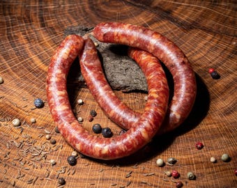 May include: Two thick, red-brown sausages are coiled together on a wooden surface. The surface is covered in small, black peppercorns and other spices.