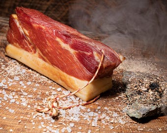 May include: A close-up of a large piece of smoked meat on a wooden surface. The meat is tied with twine and surrounded by salt crystals and wood shavings.