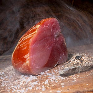 Könnte beinhalten: Ein großes Stück geräucherten Schinkens auf einer Holzoberfläche mit Salzkrusten und Holzspänen. Der Schinken ist dunkelrot und hat einen leicht orangenen Farbton. Aus dem Schinken steigt Rauch auf.