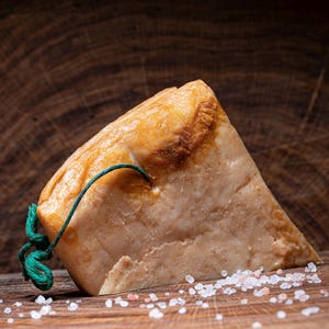 Könnte beinhalten: Ein dreieckiger Block aus hellrosa Käse mit einer braunen, leicht verbrannten Oberfläche. Der Käse ist mit einem grünen Band gebunden und liegt auf einer Holzoberfläche mit verstreuten rosa Salzkristallen.