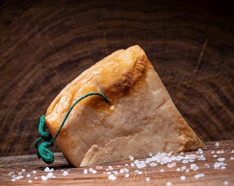 May include: A triangular block of pale pink cheese with a brown, slightly burnt surface. The cheese is tied with a green string and is sitting on a wooden surface with pink salt crystals scattered around it.