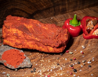 May include: A large piece of red-colored cured meat on a wooden surface with red peppers, salt, and peppercorns scattered around it. There is also a small pile of red paprika on a gray rock.