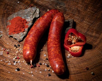 May include: Two red sausages with a red pepper half, red pepper flakes, salt, and peppercorns on a wooden surface.