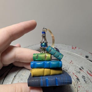 Puede incluir: Una pila en miniatura de cuatro libros en tonos azul oscuro, amarillo, azul brillante y verde, rematada con un adorno de metal plateado con una pequeña cuenta azul y un lazo de cinta dorada para colgar como dije decorativo.