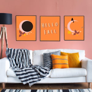 Könnte beinhalten: Drei gerahmte Drucke mit orangefarbenem Hintergrund. Der linke Druck zeigt einen Halbmond mit einem schwarzen Kreis darin. Der mittlere Druck sagt "HELLO FALL" in weißen Buchstaben. Der rechte Druck zeigt einen Halbmond mit einer schwarzen Katze, die darauf sitzt.