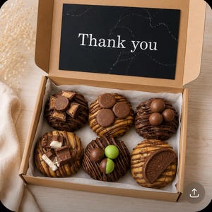 May include: A cardboard gift box containing six round cookies topped with various chocolate sweets and drizzled with chocolate. A black card inside the lid features the white text Thank you. The cookies are arranged in two rows of three on white paper.