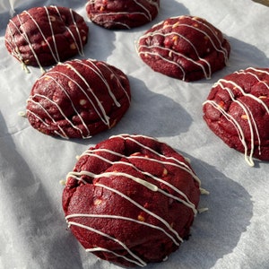 May include: Six red velvet cookies with white chocolate drizzle on a white parchment paper background.