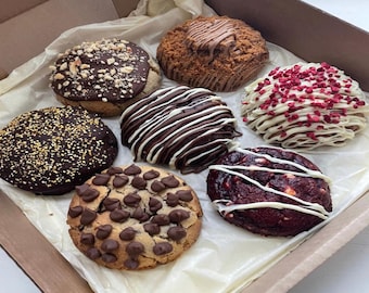 Könnte beinhalten: Eine Auswahl an Gourmet-Cookies in einer Pappschachtel. Die runden Kekse sind mit Schokoladenstückchen, Nüssen und weißem Schokoladenüberzug verziert. Einige Kekse haben eine rote Samtbasis.