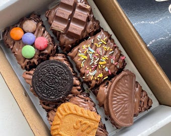 May include: A cardboard box containing six square chocolate brownies topped with various sweets. The toppings include colorful candy-coated chocolates, a chocolate bar segment, rainbow sprinkles, a dark sandwich cookie, and a chocolate orange slice.