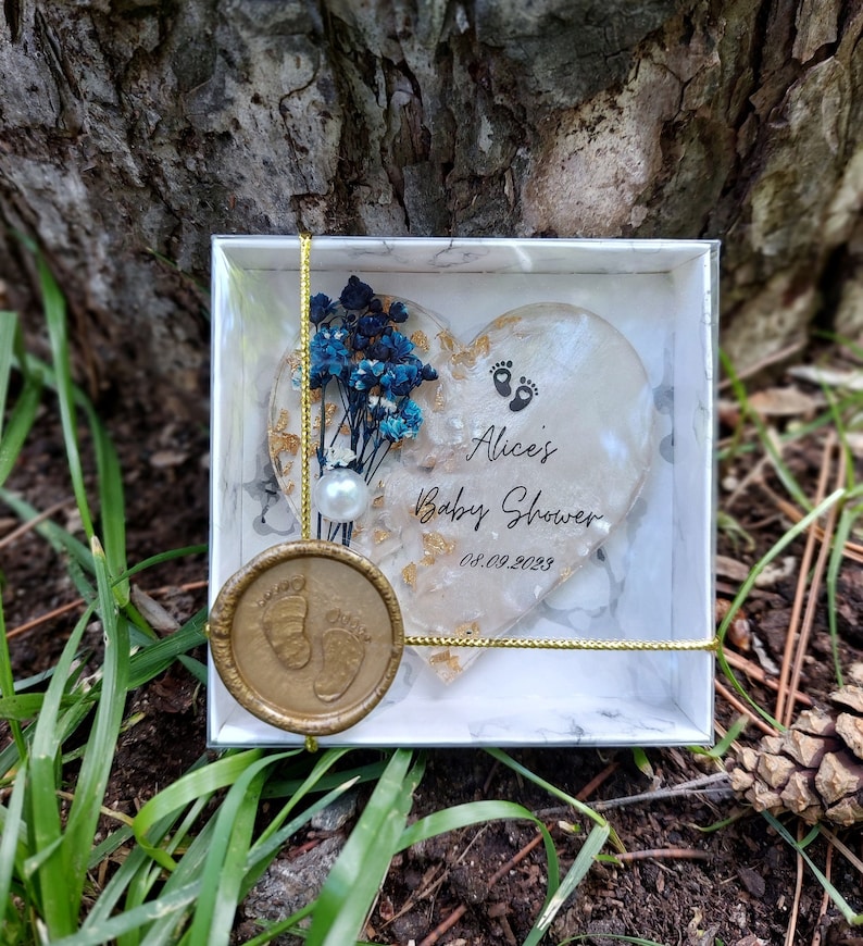 Pu&ograve; includere: Un ricordo per baby shower in una scatola bianca con un design a forma di cuore. Il cuore &egrave; decorato con fiori blu, scaglie d'oro e la scritta "Alice's Baby Shower 08.09.2023". &Egrave; attaccato un sigillo di cera dorata con impronte di piedi di bambino.