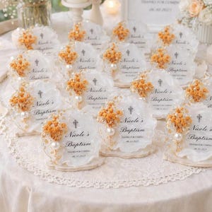Op de afbeelding: Doopbedankjes. Elk bedankje is een helder stuk met geschulpte randen, een kruis, de naam "Nicole's Baptism" en de datum. Ze zijn versierd met gedroogde oranje bloemen en een parel.