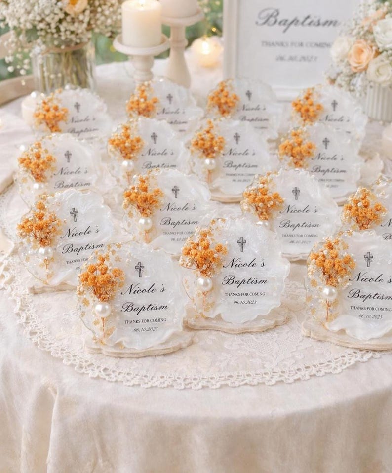 Op de afbeelding: Doopbedankjes. Elk bedankje is een helder stuk met geschulpte randen, een kruis, de naam "Nicole's Baptism" en de datum. Ze zijn versierd met gedroogde oranje bloemen en een parel.