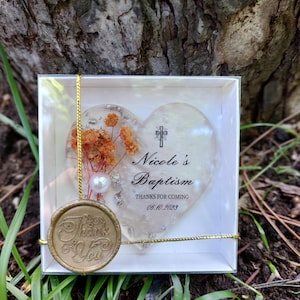 Op de afbeelding: Een hartvormig doopcadeau in een doorzichtige doos. Het hart heeft gedroogde oranje bloemen en een parel. De tekst op het hart luidt "Nicole's Doop" en "Bedankt voor uw komst 06.10.2023". Een gouden zegel zegt "Bedankt".