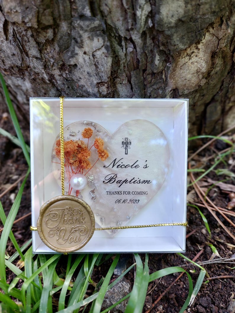 Op de afbeelding: Een hartvormig doopcadeau in een doorzichtige doos. Het hart heeft gedroogde oranje bloemen en een parel. De tekst op het hart luidt "Nicole's Doop" en "Bedankt voor uw komst 06.10.2023". Een gouden zegel zegt "Bedankt".
