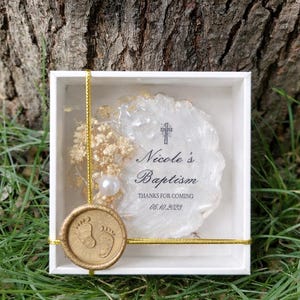 Op de afbeelding: Een witte doos met een gouden touwtje en een gouden zegel. Binnenin een wit en goud decoratief stuk met de tekst "Nicole's Baptism" en "Thanks for coming 06.10.2023". Een parel en droogbloemen zijn ook aanwezig.