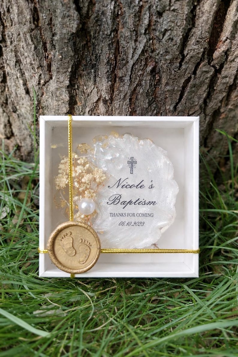Op de afbeelding: Een witte doos met een gouden touwtje en een gouden zegel. Binnenin een wit en goud decoratief stuk met de tekst "Nicole's Baptism" en "Thanks for coming 06.10.2023". Een parel en droogbloemen zijn ook aanwezig.
