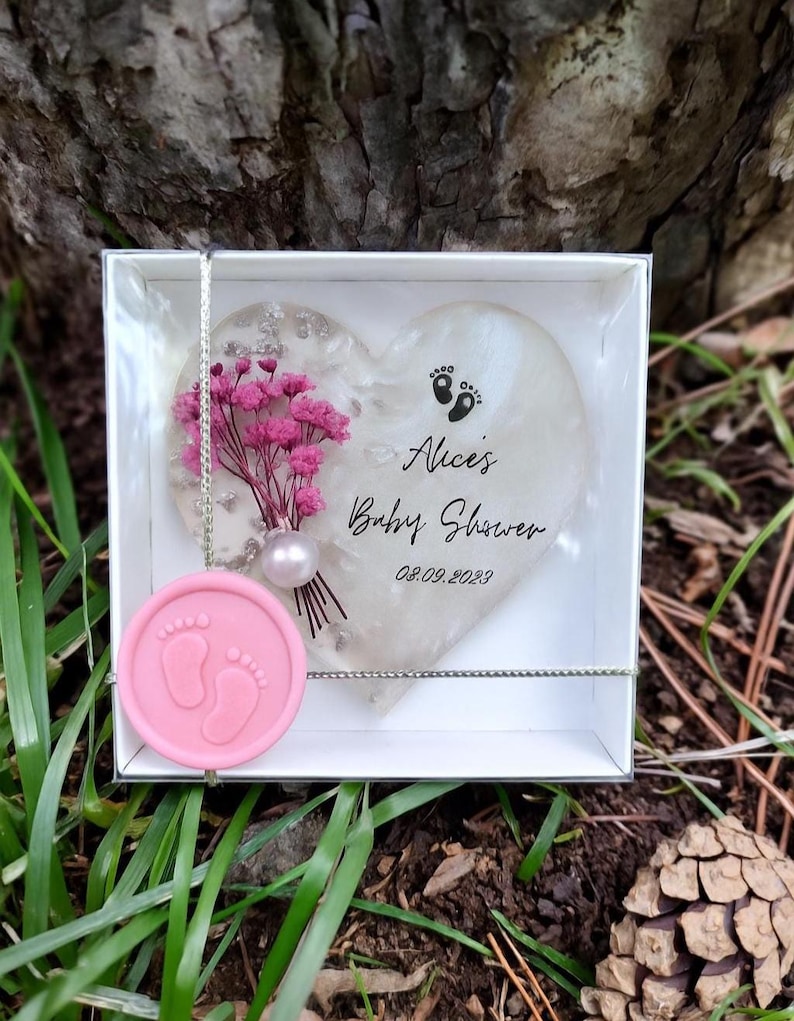 Pu&ograve; includere: Un segnaposto per baby shower con un design a forma di cuore con la scritta "Alice's Baby Shower" e la data 08.09.2023. Il cuore &egrave; decorato con fiori rosa e una perla, ed &egrave; presentato in una scatola bianca con un sigillo di cera rosa.