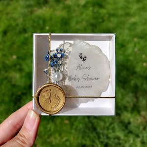 Pu&ograve; includere: Un ricordo per baby shower in una scatola bianca, con un sigillo di cera dorata con impronte di piedi di bambino. La scatola contiene un elemento decorativo bianco e argento con fiori blu, una perla e il testo "Alice's Baby Shower 08.09.2023".