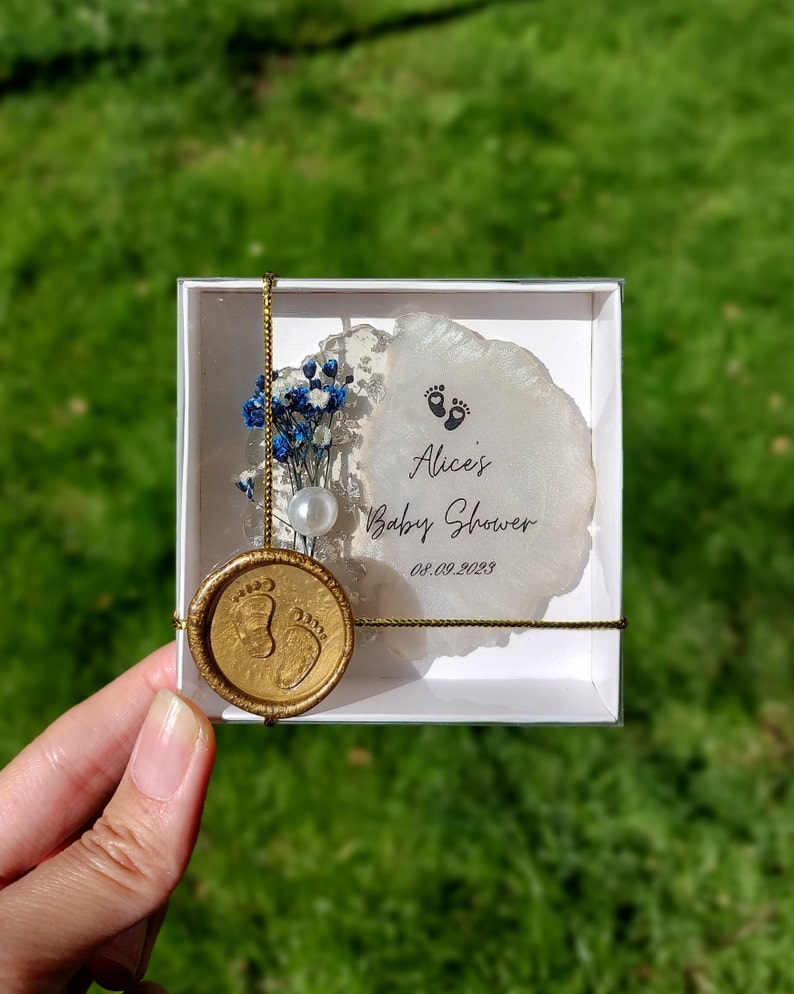 Pu&ograve; includere: Un ricordo per baby shower in una scatola bianca, con un sigillo di cera dorata con impronte di piedi di bambino. La scatola contiene un elemento decorativo bianco e argento con fiori blu, una perla e il testo "Alice's Baby Shower 08.09.2023".