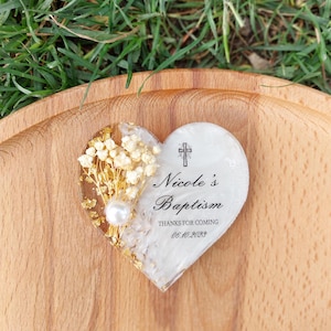 Op de afbeelding: Hartvormig doopcadeau met goudvlokken, gedroogde bloemen en een parel. De tekst "Nicole's Baptism" is afgebeeld, samen met een kruis en de datum "06.10.2023". Het cadeau ligt op een houten oppervlak.