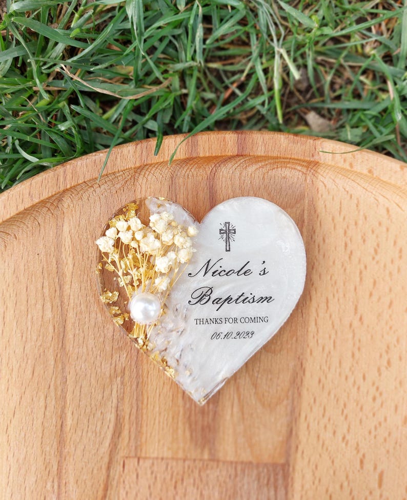 Op de afbeelding: Hartvormig doopcadeau met goudvlokken, gedroogde bloemen en een parel. De tekst "Nicole's Baptism" is afgebeeld, samen met een kruis en de datum "06.10.2023". Het cadeau ligt op een houten oppervlak.