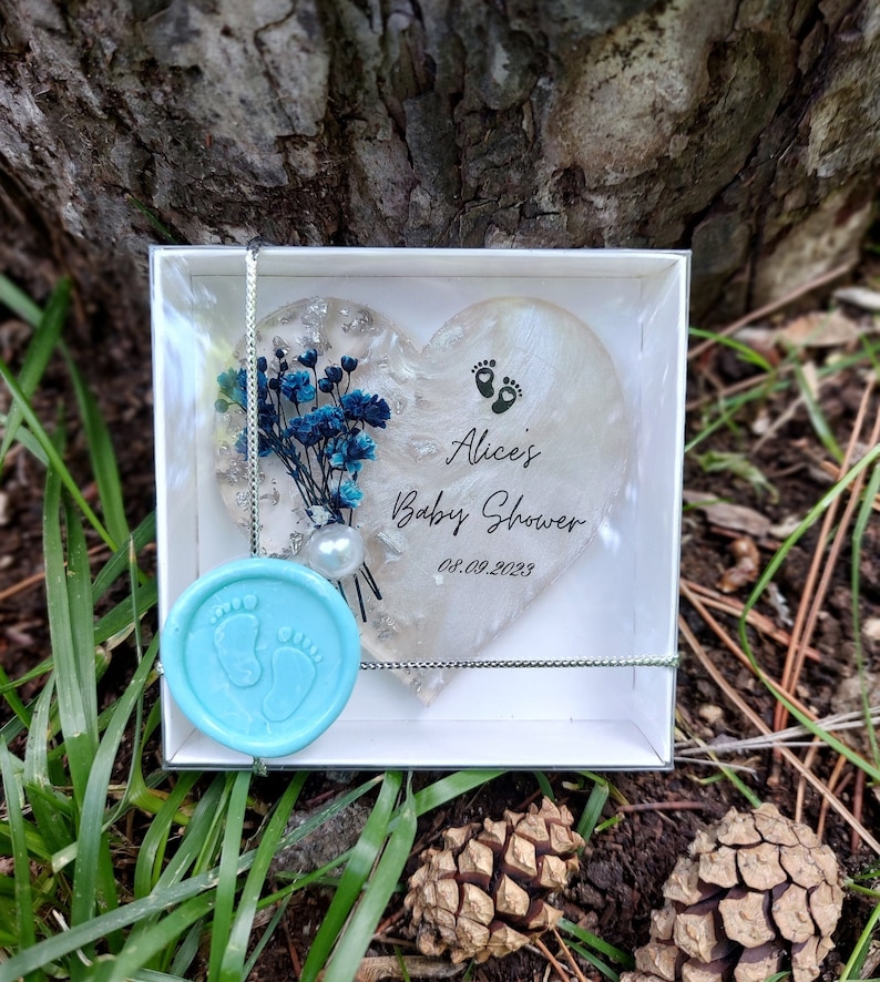 Pu&ograve; includere: Un segnaposto per baby shower a forma di cuore con la scritta "Alice's Baby Shower" e la data 08.09.2023. Il segnaposto include fiori blu, dettagli argentati e un sigillo di cera azzurra con impronte di piedi. &Egrave; presentato in una scatola trasparente.