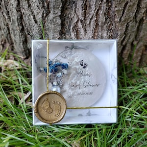 Pu&ograve; includere: Un ricordo per baby shower in una scatola bianca con un design marmorizzato. Il ricordo &egrave; un ornamento rotondo e trasparente con il testo "Alice's Baby Shower" e la data 08.09.2023. &Egrave; decorato con fiori blu e un sigillo di cera dorata.
